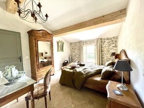 A bedroom with a bed, a desk with a chair, a dresser, a mirror, a chandelier, and a window with curtains.
