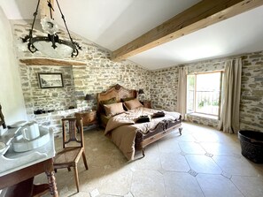 A bedroom with a stone wall, a wooden bed frame, a desk with a chair, and a window with curtains. - Gîte-Deluxe-Countryside view-Ensuite (Servies-en-Val)