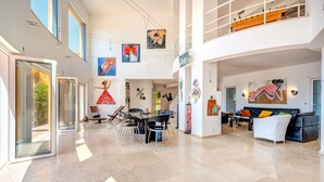 Un vaste espace de vie comprenant une table à manger, des chaises et un canapé. Les murs sont ornés d’œuvres d’art et un balcon avec une balustrade est également présent.