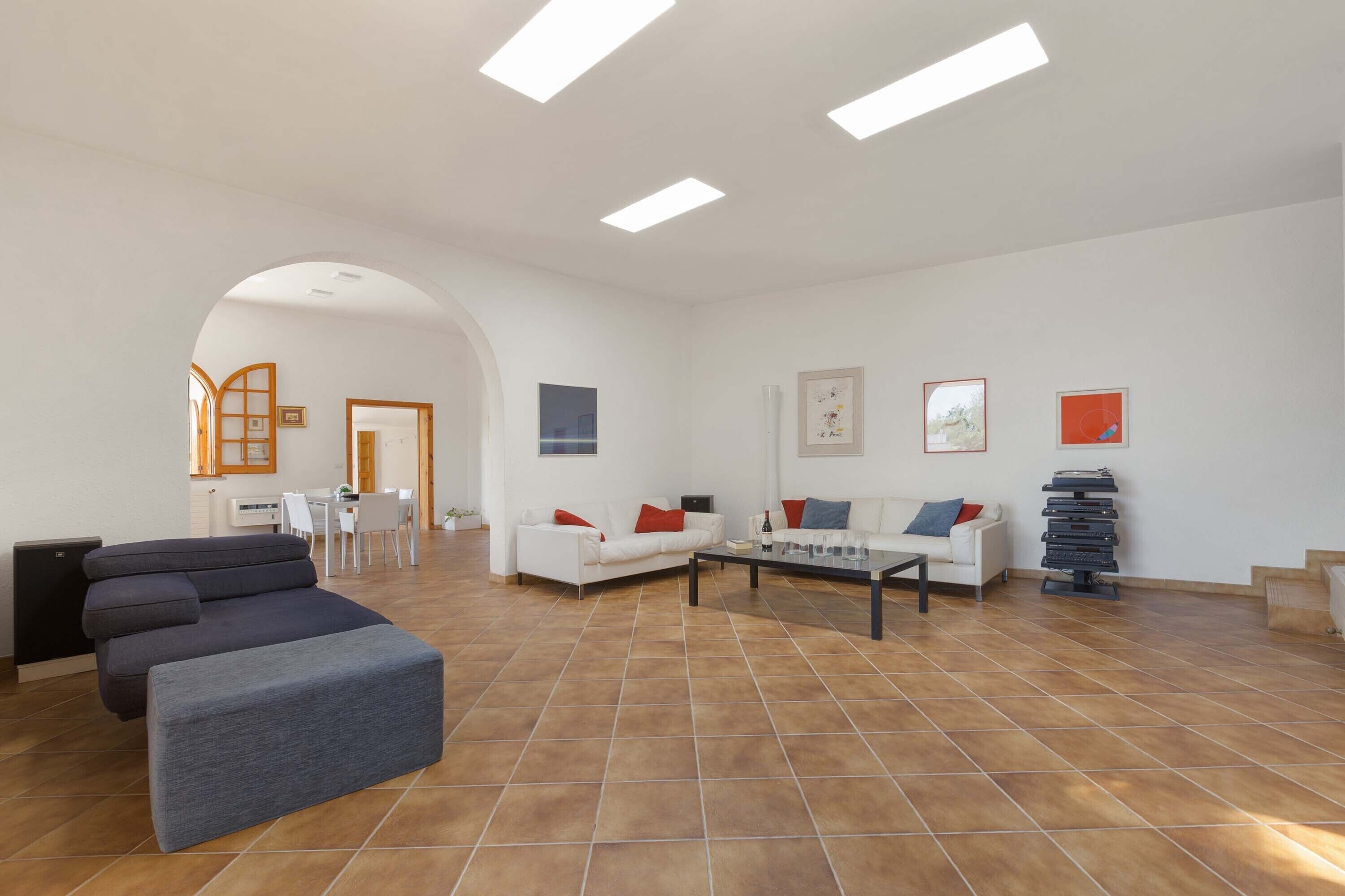 Ein geräumiges Wohnzimmer mit einem grauen Ecksofa, einem weißen Sofa und einem Couchtisch. An den Wänden befinden sich Bilder in Rahmen, und eine Treppe führt zu einer weiteren Ebene.