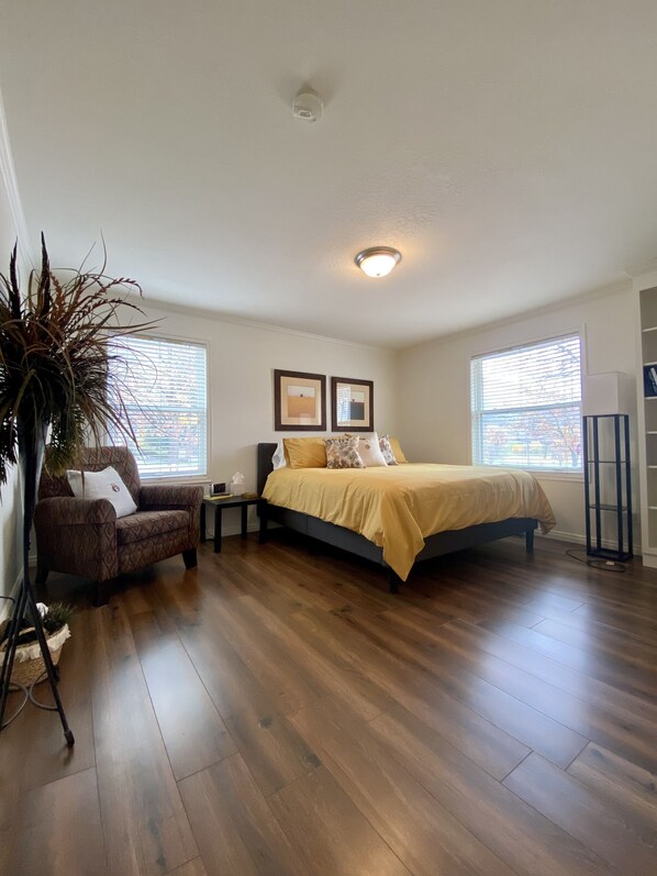 A bedroom with a bed, a chair, a nightstand, a floor lamp, and a large plant.