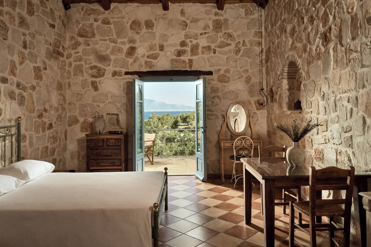 A rustic stone-walled room with a bed, a wooden desk, and a view of the outdoors.