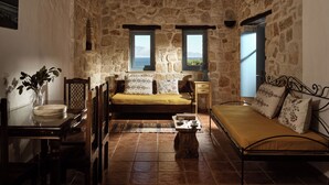 A rustic stone room with a bed, a wooden table, and a view of the sea through two windows. - Strofilia Stone Residences (Zakynthos)