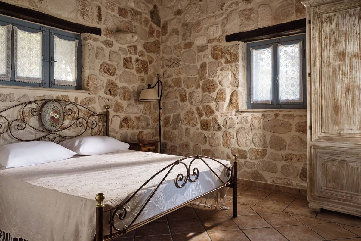 A stone-walled bedroom with a bed, two windows, a lamp, and a wardrobe.