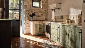 A rustic kitchen with stone walls, a wooden cabinet, a tiled backsplash, and a window with a view of greenery outside. - Strofilia Stone Residences (Zakynthos)