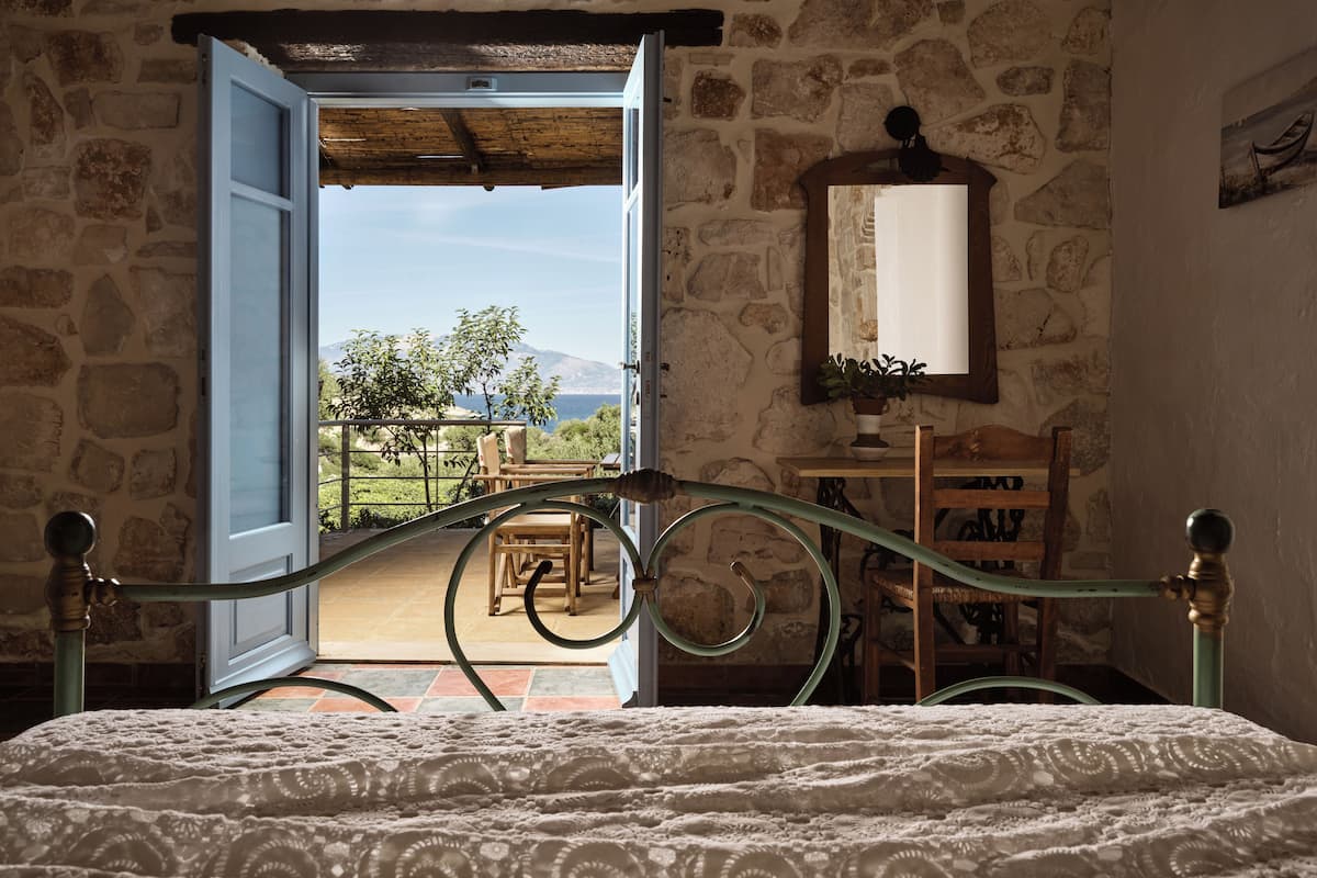 A bedroom with a stone wall, a wooden bed frame, a view through an open door to a balcony, and a rustic interior.