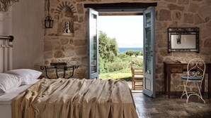 A bedroom with a stone wall, a bed with a patterned blanket, a desk with a chair, and a view of the outdoors. - Strofilia Stone Residences (Zakynthos)