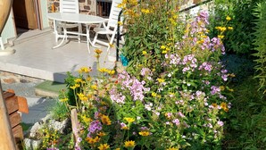 Un jardín con flores amarillas y rosas, un muro de piedra y un patio con sillas blancas.