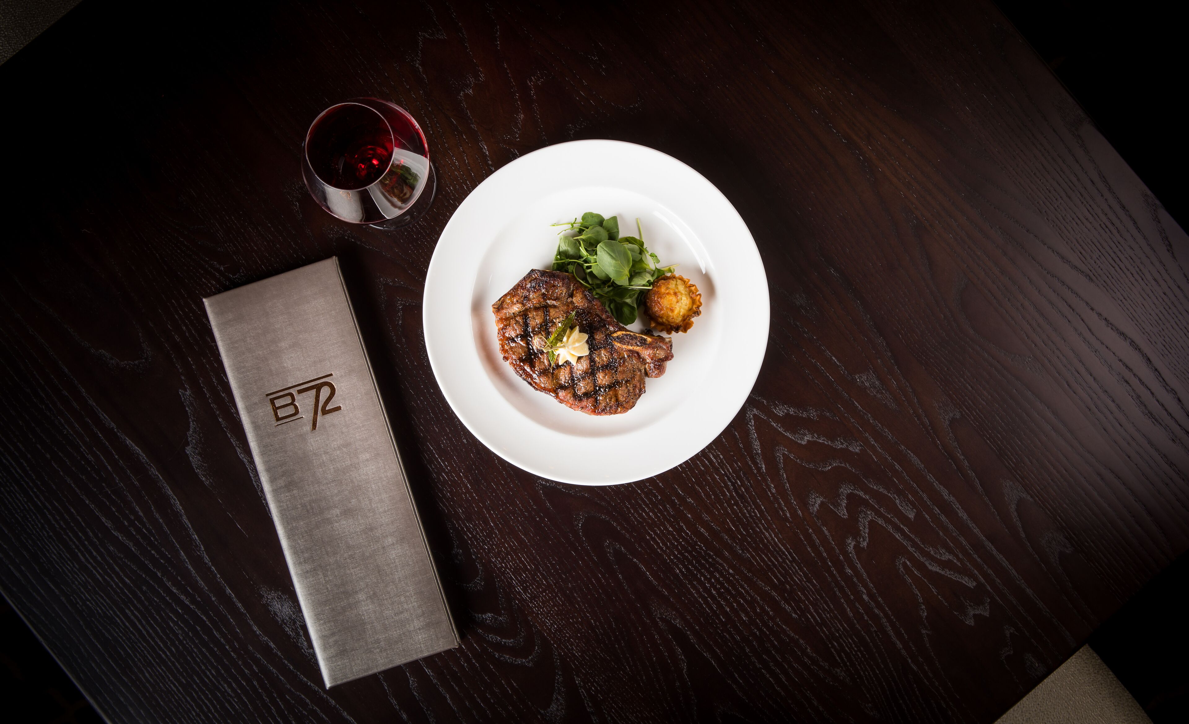 A steak with a garnish on a white plate, a glass of red wine, and a metallic napkin with a logo on a dark wooden table.