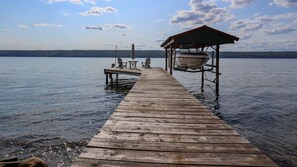 Un muelle de madera se extiende sobre aguas tranquilas, con un área de estar cubierta y un bote amarrado a la derecha.