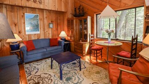 A cozy cabin interior with a wooden interior, a sofa, a coffee table, a dining table, and a window with a view of trees.