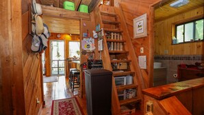 A rustic wooden cabin interior with a kitchen area, wooden shelves, and a ladder leading to a loft space.
