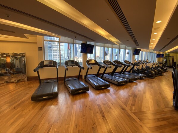 A gym with treadmills aligned along the wall, large windows, and a wooden floor.