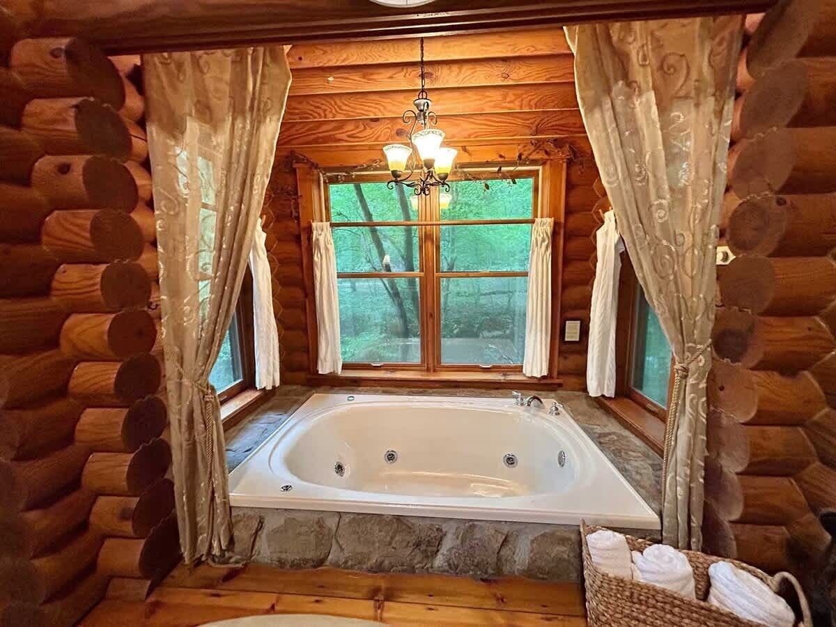 A log cabin interior with a stone floor, a large bathtub, and a window with curtains.