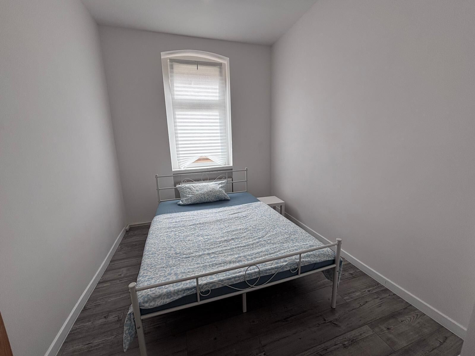 A small, single-bed room with a window, a bedside table, and a patterned bedspread.