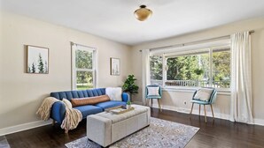 Un salon avec un canapé bleu, une petite table et un fauteuil. Il y a deux fenêtres avec des rideaux, un luminaire au plafond et des tableaux encadrés accrochés aux murs.