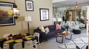 Un salon moderne avec un canapé foncé, des chaises blanches, une table basse et un coin repas agrémenté d’un saladier rouge et de verres.