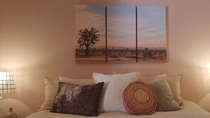Een slaapkamer met een groot bed, meerdere kussens, twee bedlampjes en een schilderij van een woestijnlandschap aan de muur.
