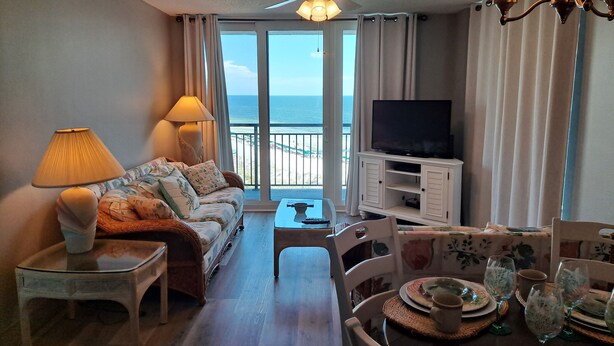 Ein Wohnzimmer mit einer Couch, einem Fernseher, einem Esstisch gedeckt mit Tellern und Gläsern sowie Blick auf den Ozean durch große Fenster.