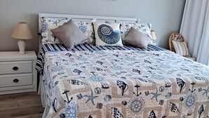 Ein ordentlich bezogenes Bett mit einer marineblauen Tagesdecke, einer Nachttischlampe und einem Gemälde mit Strandmotiv.
