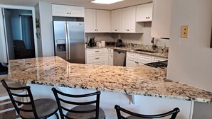 Eine Küche mit weißen Schränken, einem Edelstahl-Kühlschrank, Granit-Arbeitsplatten und einem Essbereich mit zwei Stühlen.