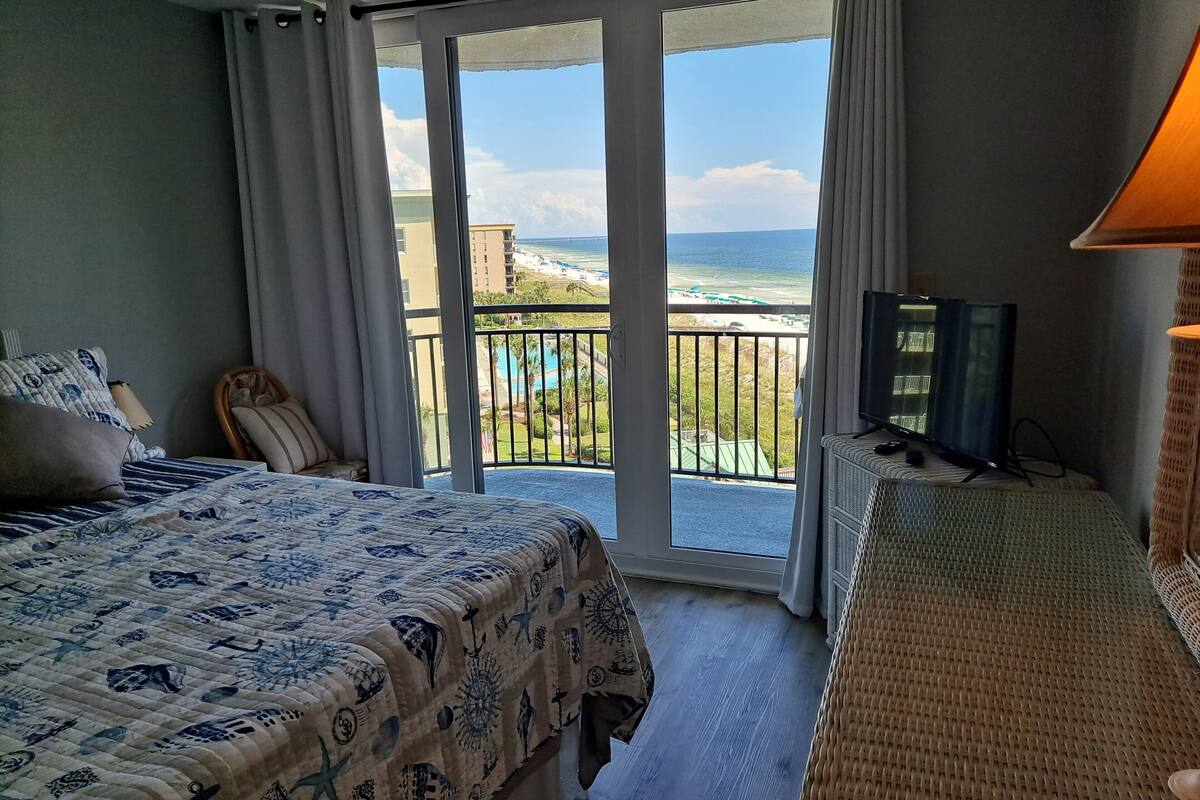 Una habitación de hotel con cama, televisión, balcón con vista al océano y piscina.