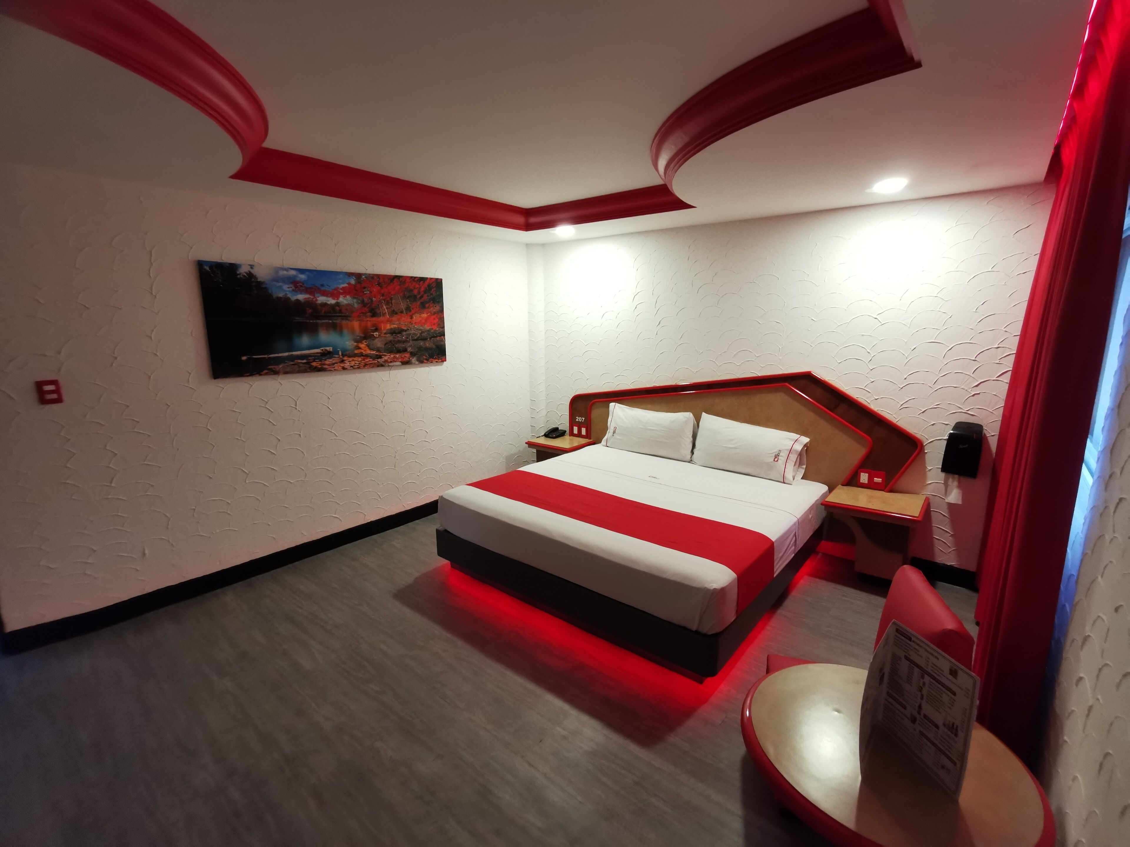 A hotel room with a bed, a nightstand, a red accent wall, a painting, and a red-lit bedside table.