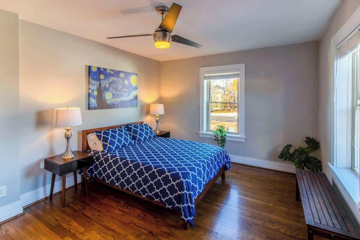 Un dormitorio con una cama, dos mesitas de noche, un ventilador de techo, una ventana y un cuadro en la pared.