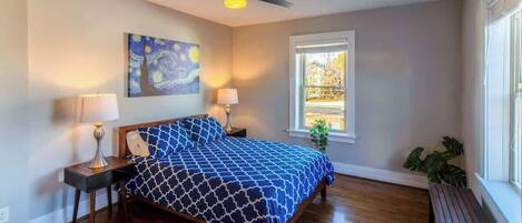 Une chambre à coucher avec un lit, deux tables de chevet, un ventilateur de plafond, une fenêtre et un tableau au mur.