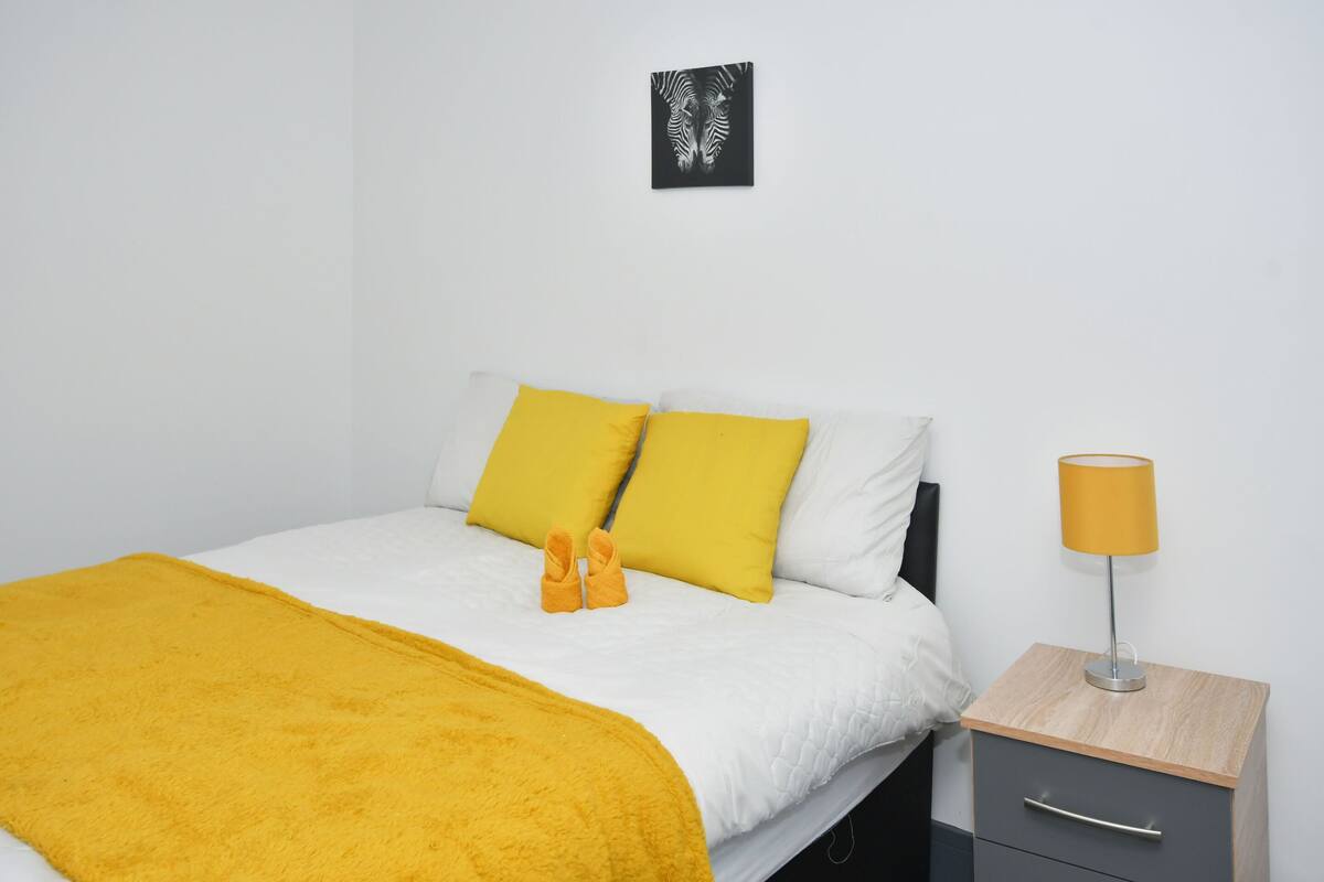 Una cama bien hecha con almohadillas amarillas y una colcha, una mesita de noche con una lámpara y una obra de arte en la pared.