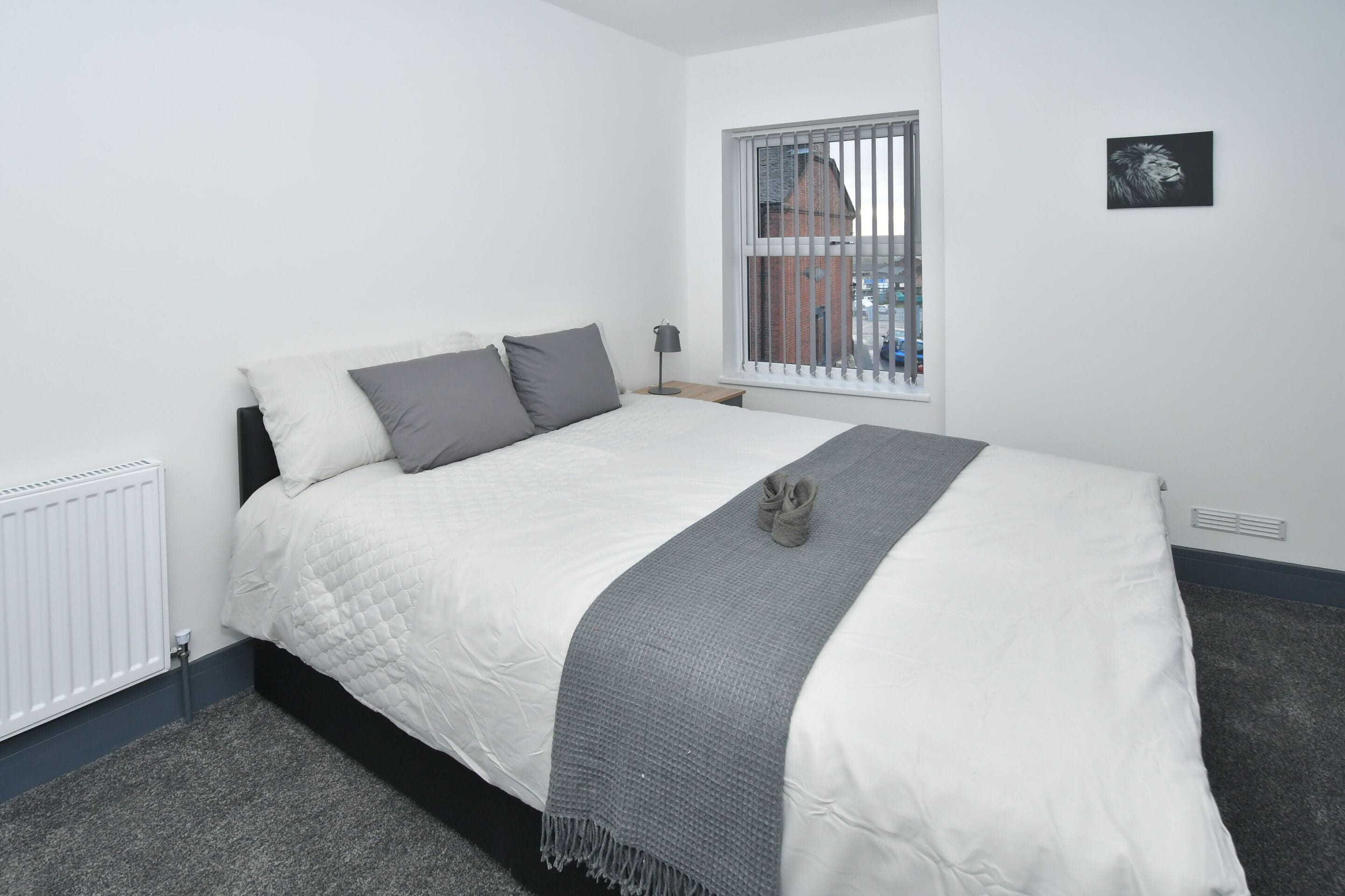 Un lit bien fait, avec une couette blanche et des oreillers gris, un plaid gris et une paire de chaussons posés sur le lit. Il y a une fenêtre avec des stores blancs, un radiateur et un petit tableau accroché au mur.