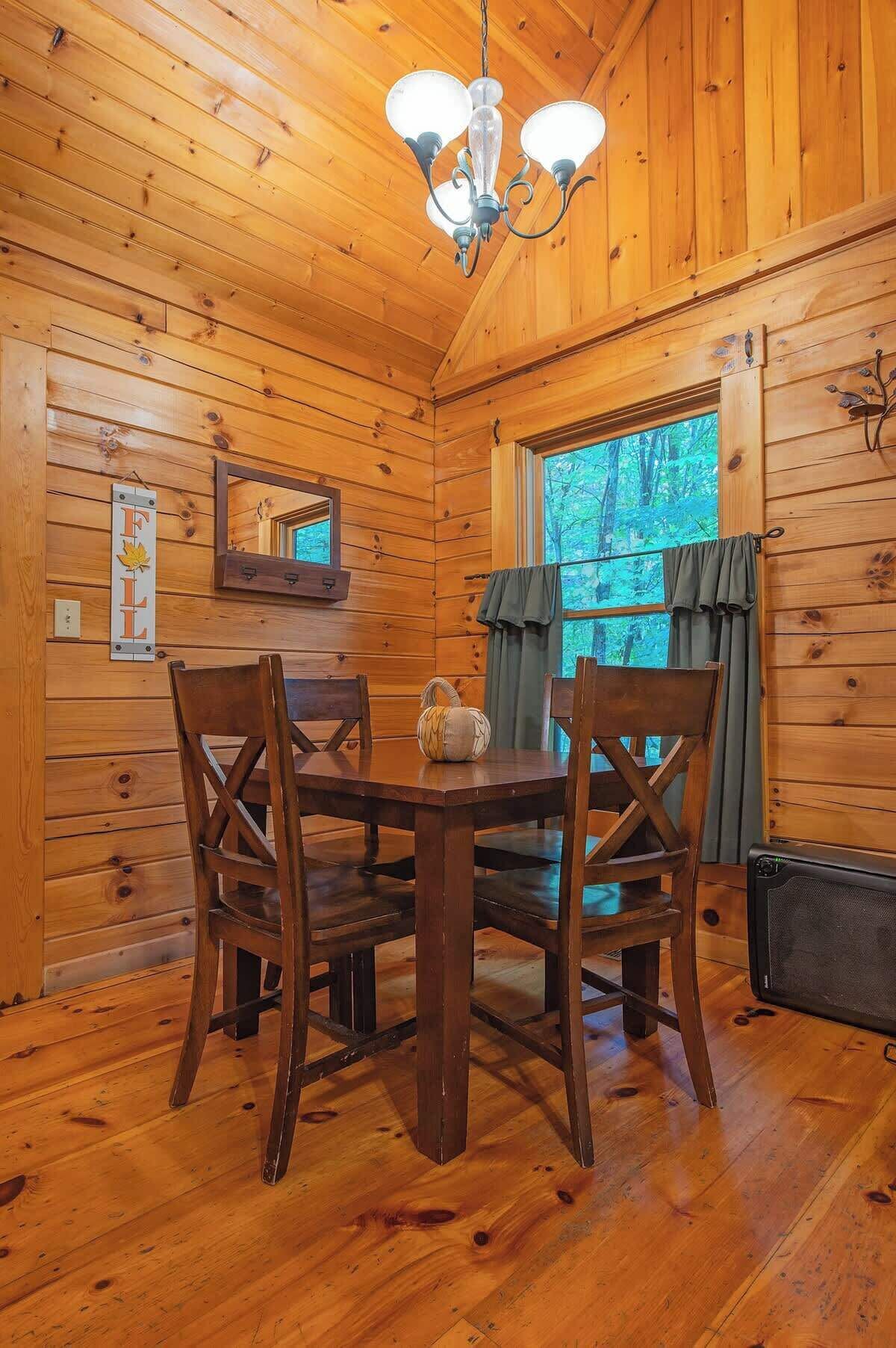Interno di una baita di legno con un tavolo da pranzo, sedie, un lampadario, una finestra con tende e uno specchio a parete.