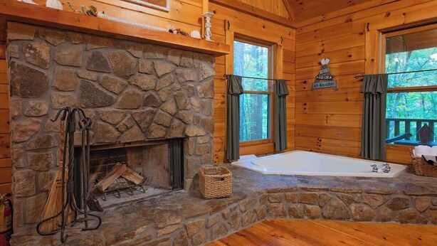 A stone fireplace with logs, a bathtub, and a window with a view of the forest. - Cherokee private cabin in Riverside Resort (Benton)