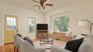 A living room with a ceiling fan, a TV, a wooden cabinet, and a window with a view of trees.