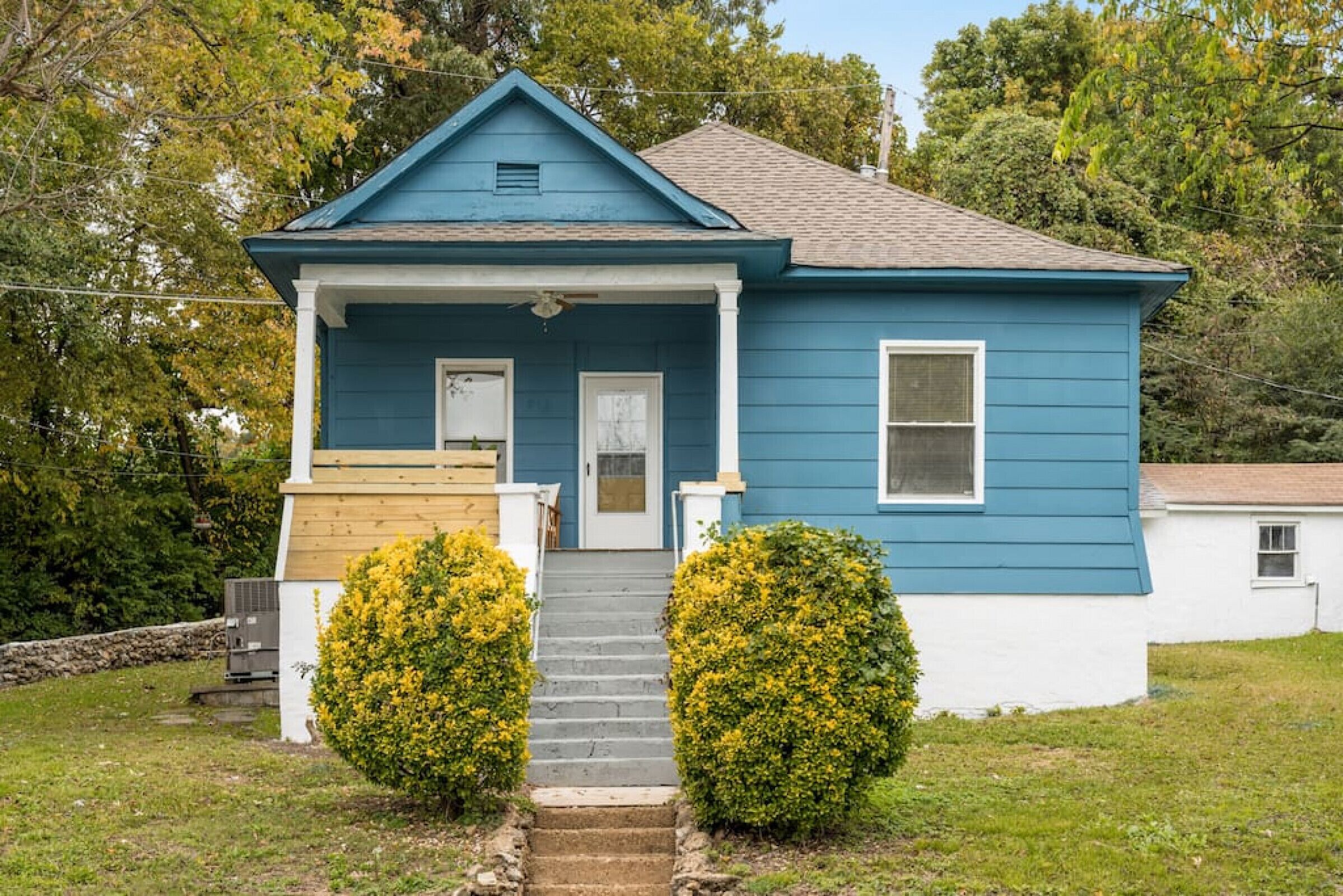 Une maison à deux étages, à l’extérieur bleu, avec un porche à l’avant et un petit jardin devant, agrémenté de deux arbustes.