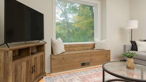 A living room with a television on a wooden stand, a bench with cushions, a glass coffee table, and a view of trees through a window.