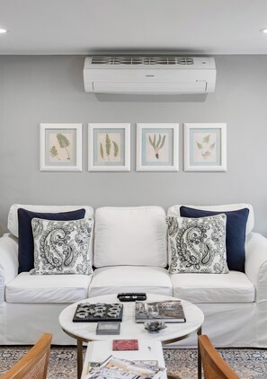Un salon avec un canapé blanc, deux coussins décoratifs à motifs, une table basse ronde sur laquelle sont posés des livres et une télécommande, et un appareil de climatisation installé au-dessus.