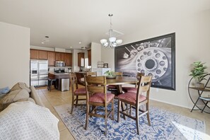 A modern kitchen with stainless steel appliances, a dining area with wooden chairs and a large painting, and a living room with a sofa and a patterned rug.