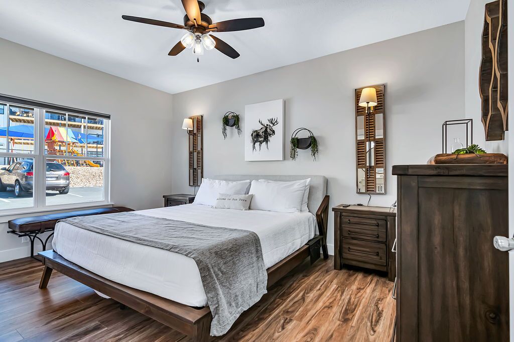 A bedroom with a bed, bedside tables, a ceiling fan, a window with a view of a playground, and wall-mounted decor.