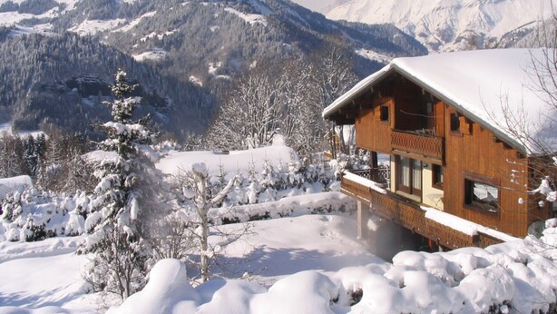 A wooden chalet with a balcony, surrounded by snow-covered trees and mountains in the background. - GITE DANS CHALET SAVOYARD, 4 PERS, au pied des pistes, calme, vue, expo sud (NOTRE-DAME-DE-BELLECOMBE (73590))