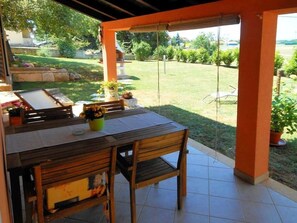 Eine Terrasse im Freien mit einem Holztisch und Stühlen, umgeben von orangefarbenen Säulen und Blick auf eine Rasenfläche.