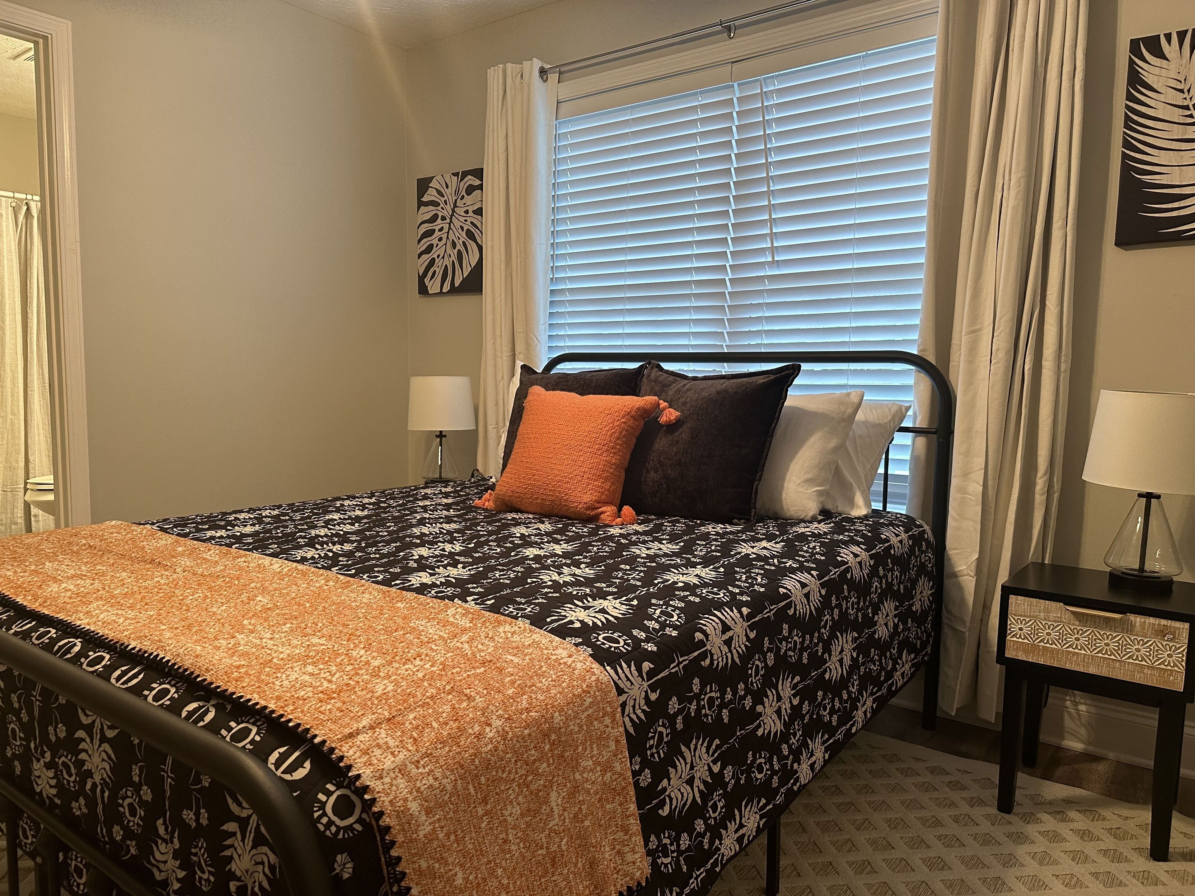 Une chambre à coucher bien rangée, avec un lit, deux tables de chevet, une fenêtre avec des stores et une œuvre d’art accrochée au mur.