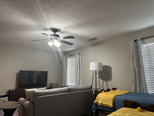 Una habitación de hotel con ventilador de techo, televisor de pantalla plana, sofá, cama con manta amarilla y lámpara de pie.