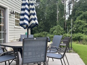 En uteplats med ett runt bord och sex stolar, ett randigt parasoll och ett hus i bakgrunden.