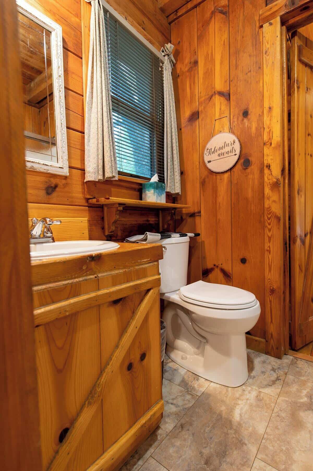 Un baño estilo cabaña de madera con lavamanos, inodoro y ventana con cortinas.