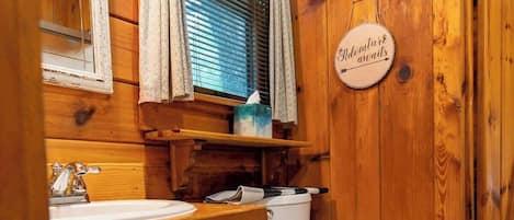 Un baño estilo cabaña de madera con lavamanos, inodoro y ventana con cortinas.