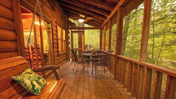 Une cabane en bois avec une véranda, une table à manger, des chaises et une balançoire suspendue.