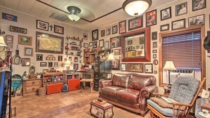 A vintage-styled living room with a brown leather sofa, a wooden armchair, and a coffee table. The room is decorated with numerous framed pictures and memorabilia on the walls.
