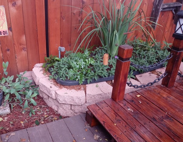 A wooden deck with a stone planter bed containing various plants, a small wooden bench, and a chain-link fence.