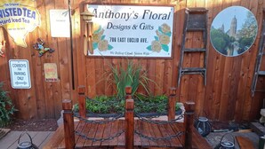 A wooden bench with a sign for Anthony's Floral Designs & Gifts, surrounded by plants and lanterns.